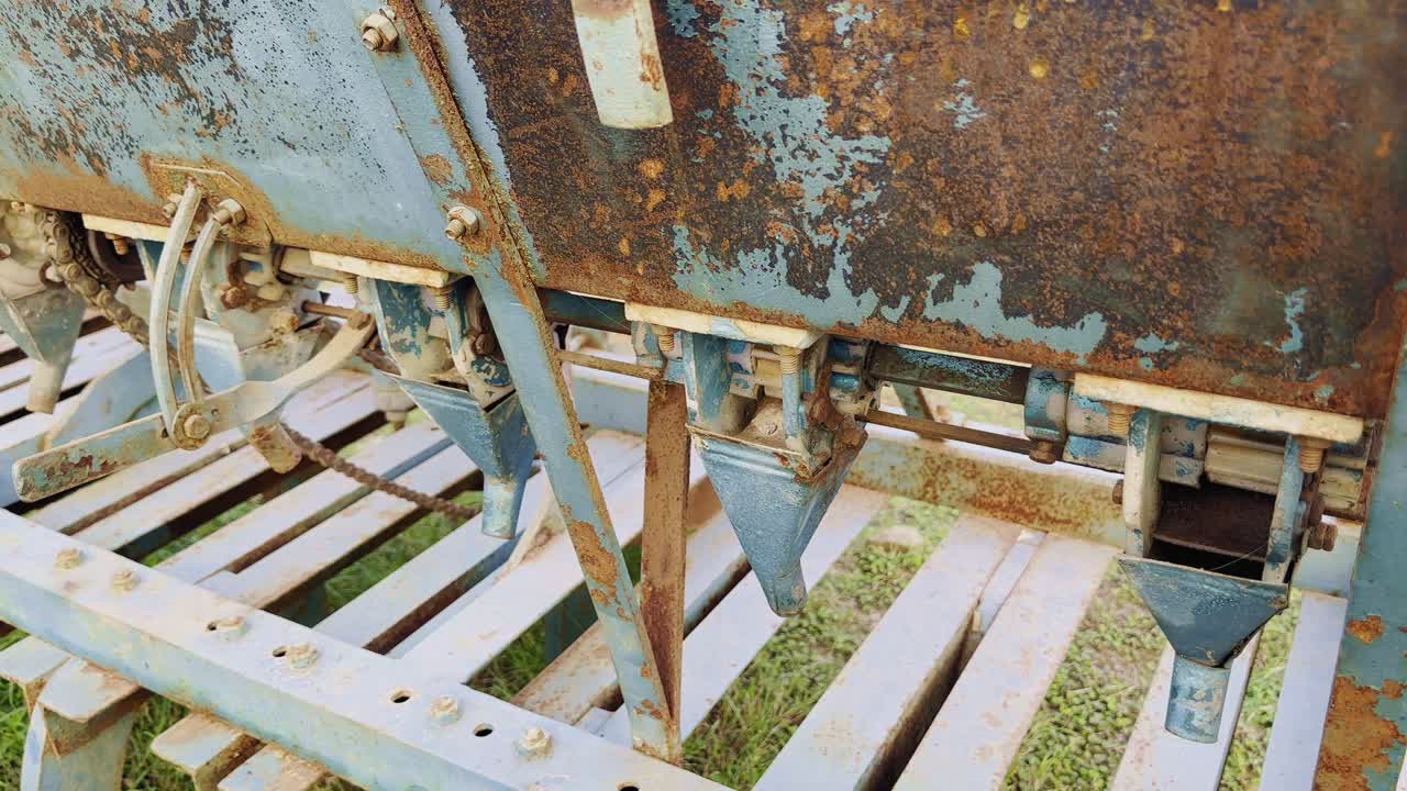 The camera moves in a circular motion around an old rusty blue seed drill machine, highlighting its worn metal body, mechanical levers, and rusted seed outlets,