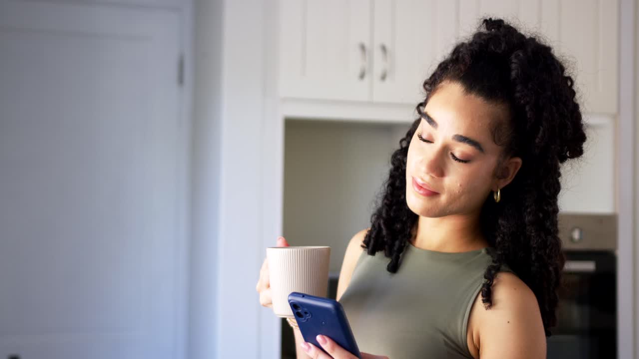 Mujer mirando de reojo una taza, acercando un smartphone azul y leyendo la pantalla en la cocina para revisar un mensaje.