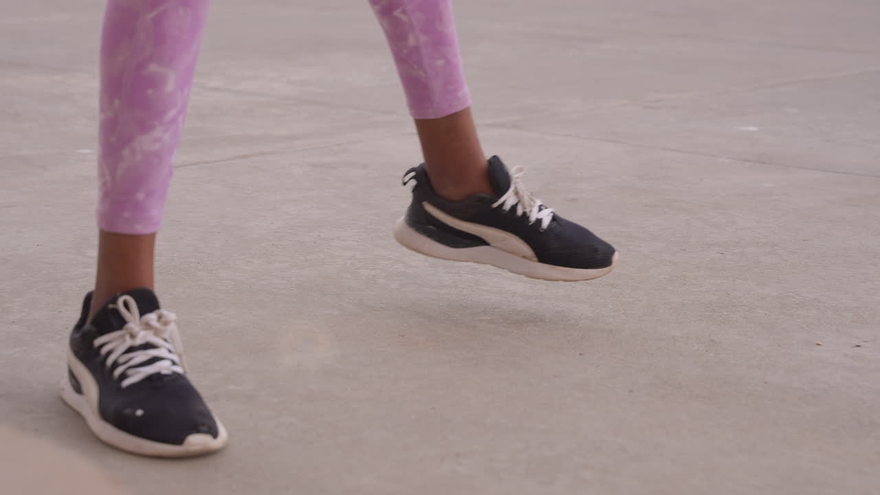 Primo piano dei piedi di una donna nera con sneakers che eseguono veloci saltelli laterali sul cemento, indossando leggings viola, ruoli di istruttrice di fitness e studentessa di danza suggeriti, atmosfera energica di studio urbano.
