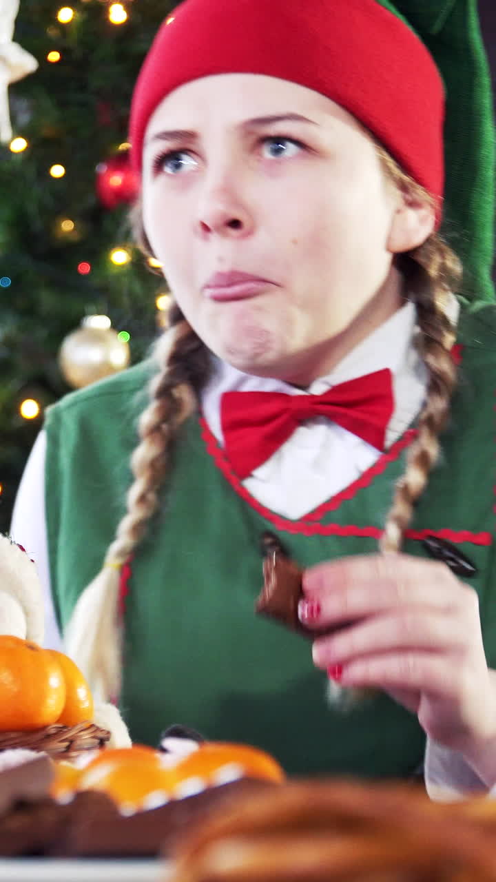 Tricky female elf sitting at table on Christmas tree background. Girl in elf suit is taking delicious food on table. Vertical video