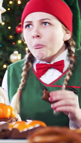 tricky female elf sitting at table on christmas tree background. girl in elf suit is taking delicious food on table. vertical video