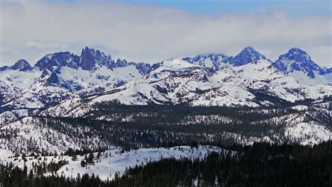 banner peak minaret summit pass mammoth mountains vista parallax winter aerial drone california group jagged towering snow covered peaks sunny blue sky inyo forest sierra nevada valley backwards