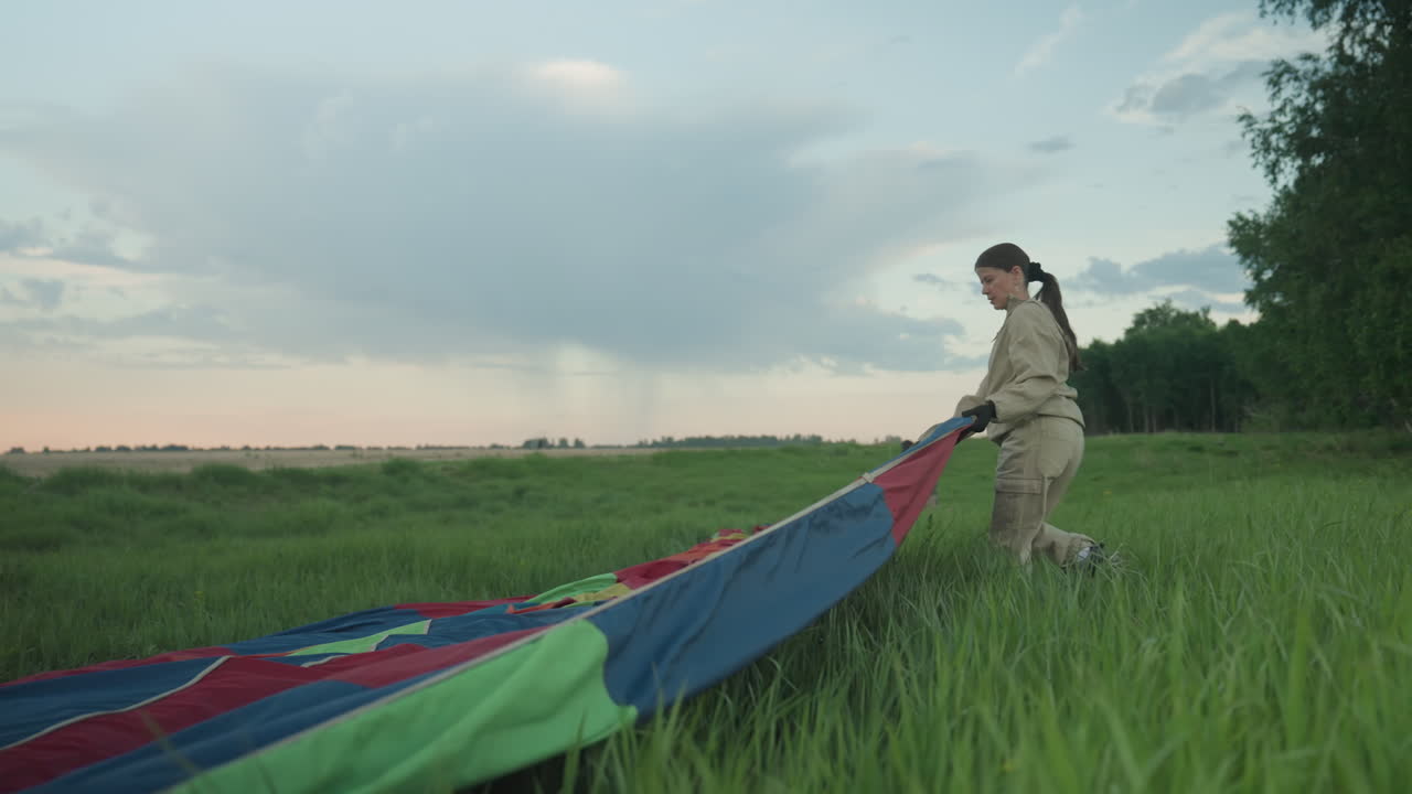 couple wearing matching khaki jackets drag multicolored hot air balloon canopy through tall grass at dusk across open field under pastel sunset sky with distant tree line during preflight setup