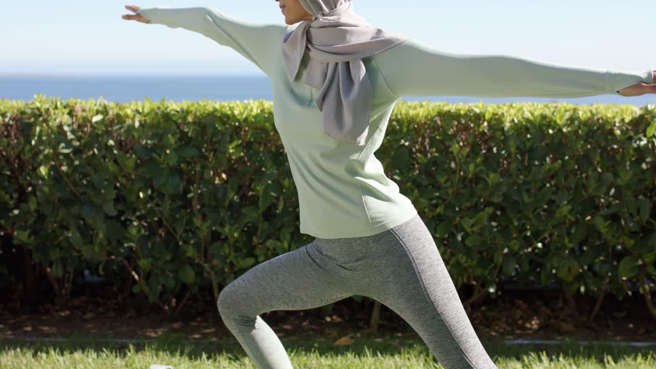 Woman in headscarf stepping onto mat on lawn, bottle nearby, moving into warrior pose for yoga
