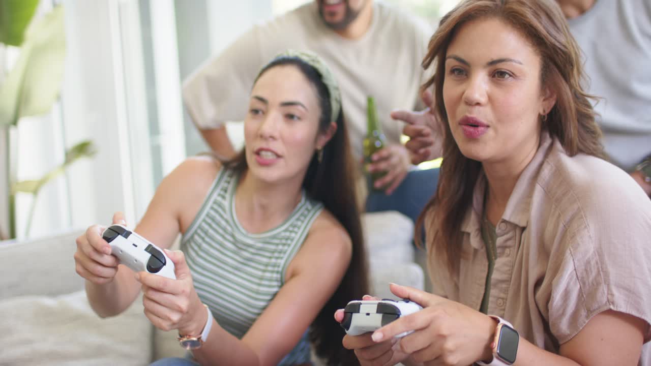 Amici che osservano due donne adulte giocare mentre il gioco inizia, stringendo controller bianchi sul divano.