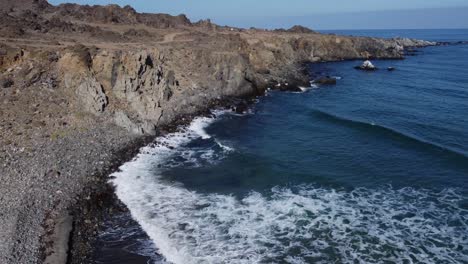 Flyover-rocky-ocean-beach-to-dirt-road-on-rock-cliffs-above-in-Chile