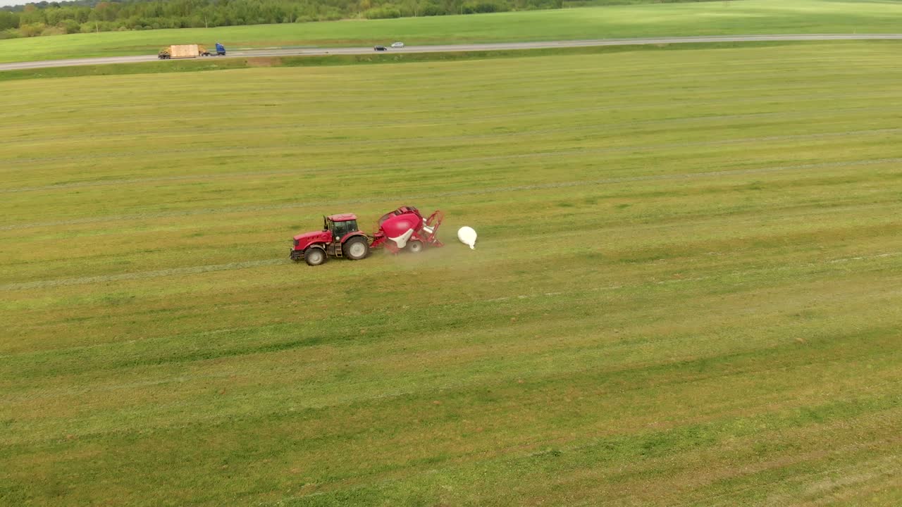 un tractor rojo recoge heno y lo presiona en balas blancas usando una balera, disparada desde arriba