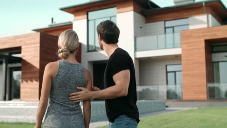 Handsome-man-closing-woman-eyes-near-house.-Surprised-woman-standing-near-villa