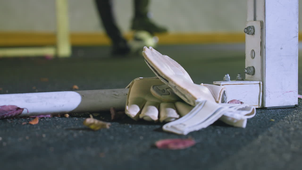 Close-up of white goalkeeper gloves resting near goalpost on dark turf surrounded by fallen leaves, with blurred player balancing soccer ball underfoot in background