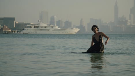 beautiful woman wearing black dress playing and dancing in the water of the sea with luxury ship in background