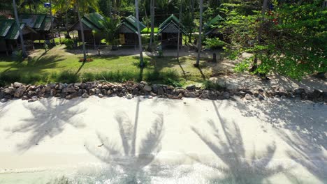 Strandhütten-Unter-Hohen-Palmen-Beschatten-Den-Weißen-Sand,-Der-Vom-Ruhigen,-Klaren-Wasser-Der-Türkisfarbenen-Lagune-In-Thailand-Umspült-Wird