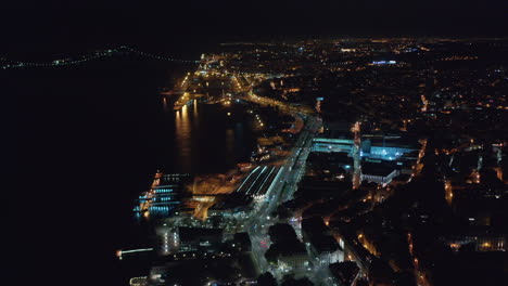 Aerial-night-view-of-urban-city-center-of-Lisbon-with-bright-lights-on-houses,-Ponte-25-de-Abril-bridge-and-coastal-traffic-by-the-sea