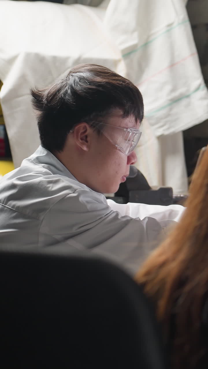 자동차 작업실에 앉아있는 실험실 기술자, 구성 요소를 작업하고, 도구와 장치를 사용하여, 조직된 장비, 형광 조명 및 기계적 도구로 둘러싸여 있습니다.