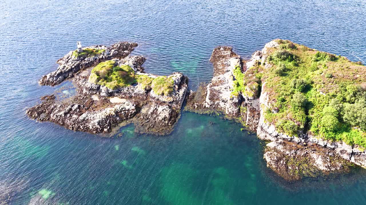 Drone glides above rocky island with lighthouse, clear coastal waters, and lush greenery in sunlight
