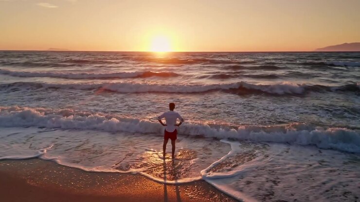Young man with white shirt walking alone along the beach enjoying the beautiful sunset. waves coming to the shore. Sun is setting. Warm dusk sky. Golden hour. Summer holidays vibes.