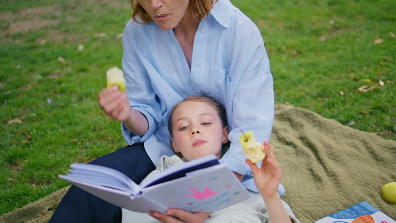 Picnic kid eating apple lying mom knees at lawn closeup. Woman reading book
