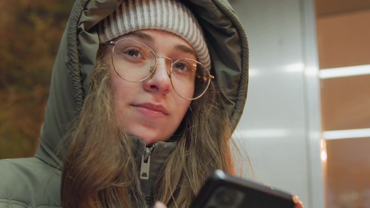 White woman in hood checking phone, pensive commuter at indoor station, wearing beanie and round glasses, scrolling messages under warm neon lights, closeup portrait with soft bokeh and film grain