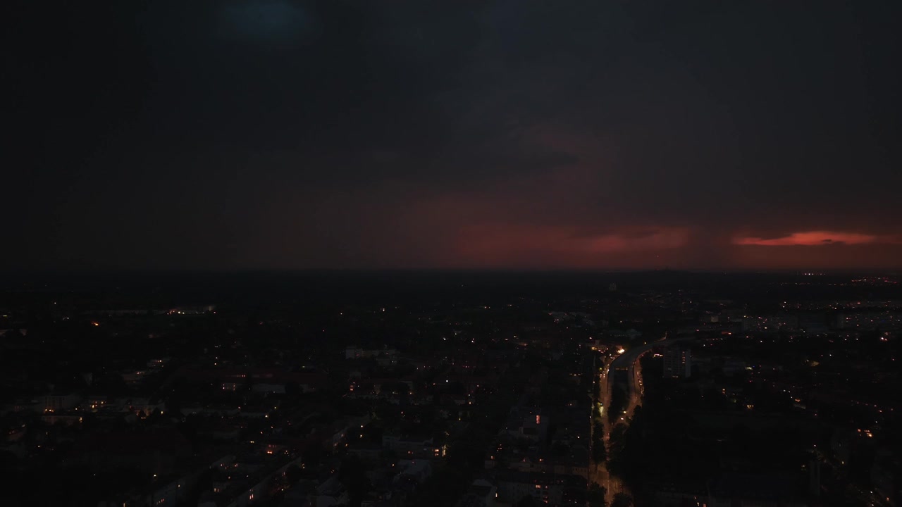 Blesky blikající nad městem v noci, osvětlující mraky a panorama s auty jedoucími po ulici během bouřky. Dramatický letecký pohled, statické panorama z vznášejícího se dronu.