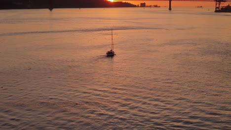 aerial establishing dolly tilt up follows sailboat on tagus river with sunset between suspension bridge