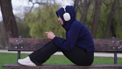 woman listen music on headphones and play game on smartphone, sit on park bench