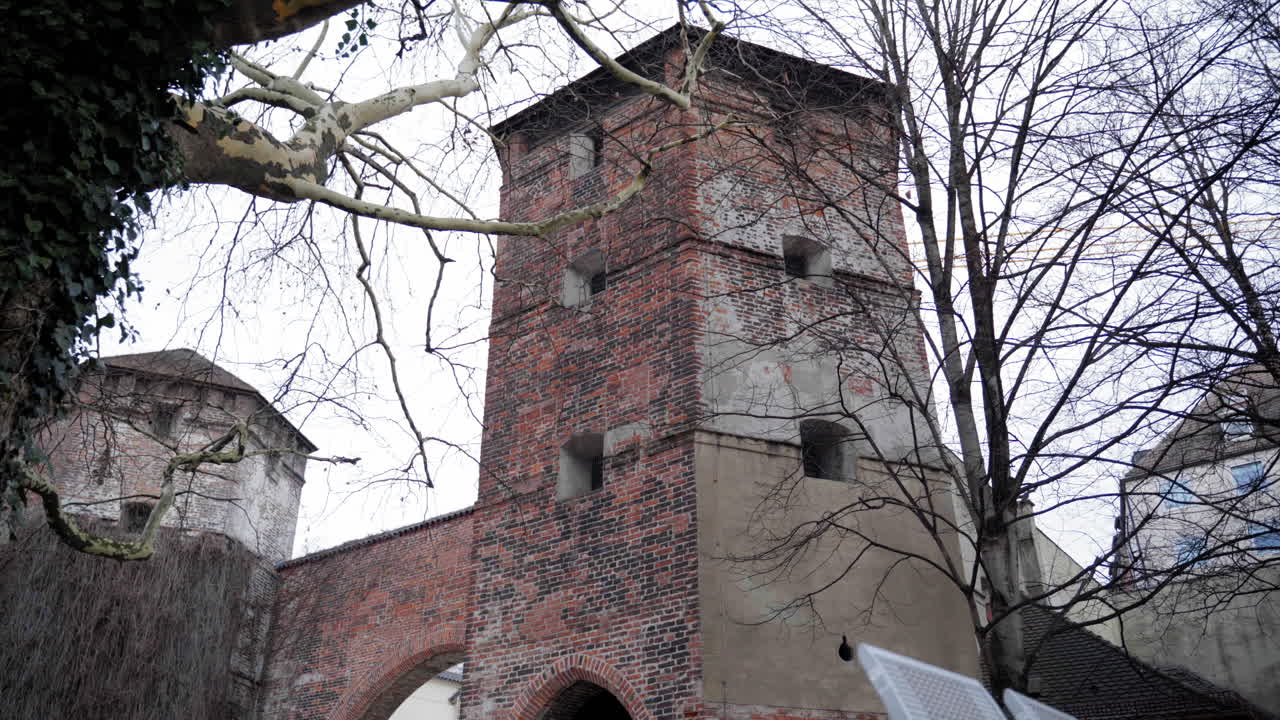 Historic medieval brick tower in Munich, Germany, part of the old city fortifications. Bare trees and an overcast sky create a winter atmosphere, highlighting the aged architecture.