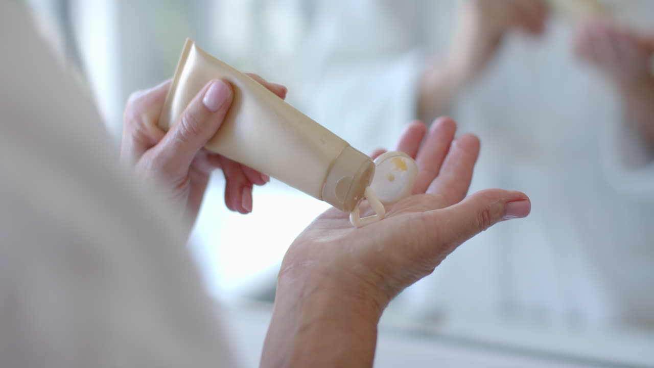 Applying lotion, mature woman squeezing cream from tube onto hand in bathroom