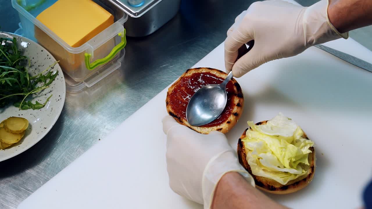 Lo chef stende la salsa rossa con un cucchiaio su un panino. Il cuoco spalma la salsa su metà panino da hamburger grigliato sul tavolo della cucina. Preparazione di fast food. Vista dall'alto
