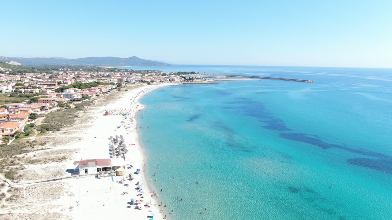 Questo video avanza lungo una costa di sabbia bianca e blu, mostrando dettagli di nuotatori, auto in transito e una città sullo sfondo.