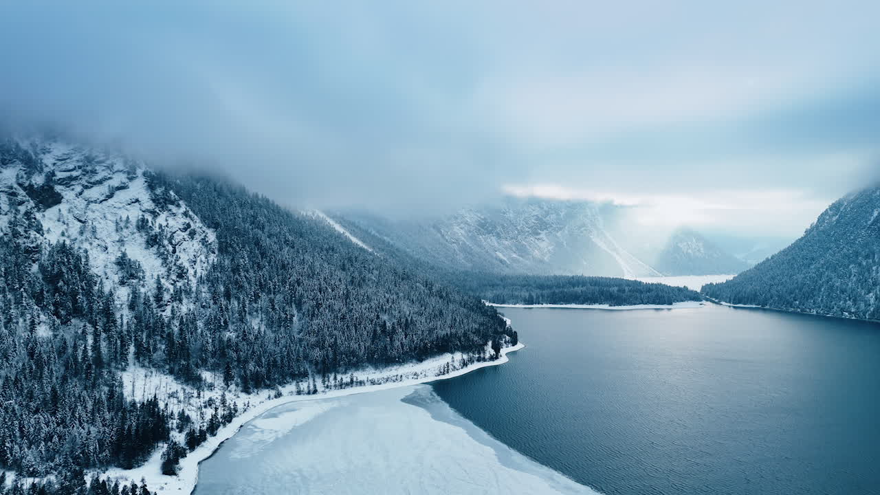 Terrific mighty mountains covered with pine tree woods. Ice covers the waterscape at the lakefront. Dramatic grey cloudscape shading the sky. Aerial view.