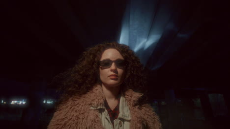 Young Woman in Stylish Sunglasses and Fluffy Jacket Posing in Dark Underpass