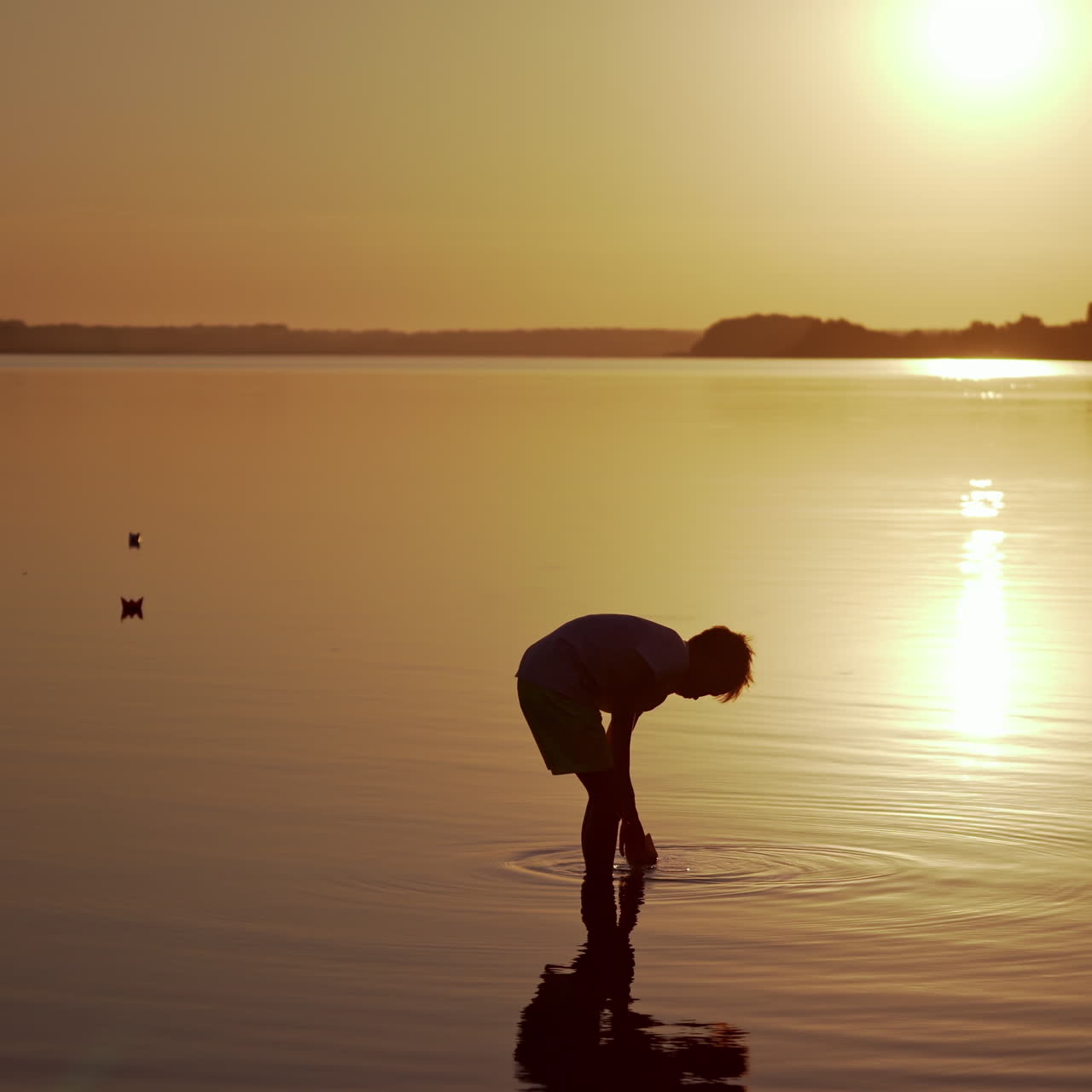 Child in water at sunset. Silhouette of a boy putting paper boat on water in the evening. Kid holding origami ship on the lake.