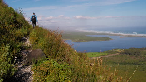 man with drone backpack hiking a track on the side of a steep mountain-side with breathtaking views of lagoon and white sand beach coastline in distance