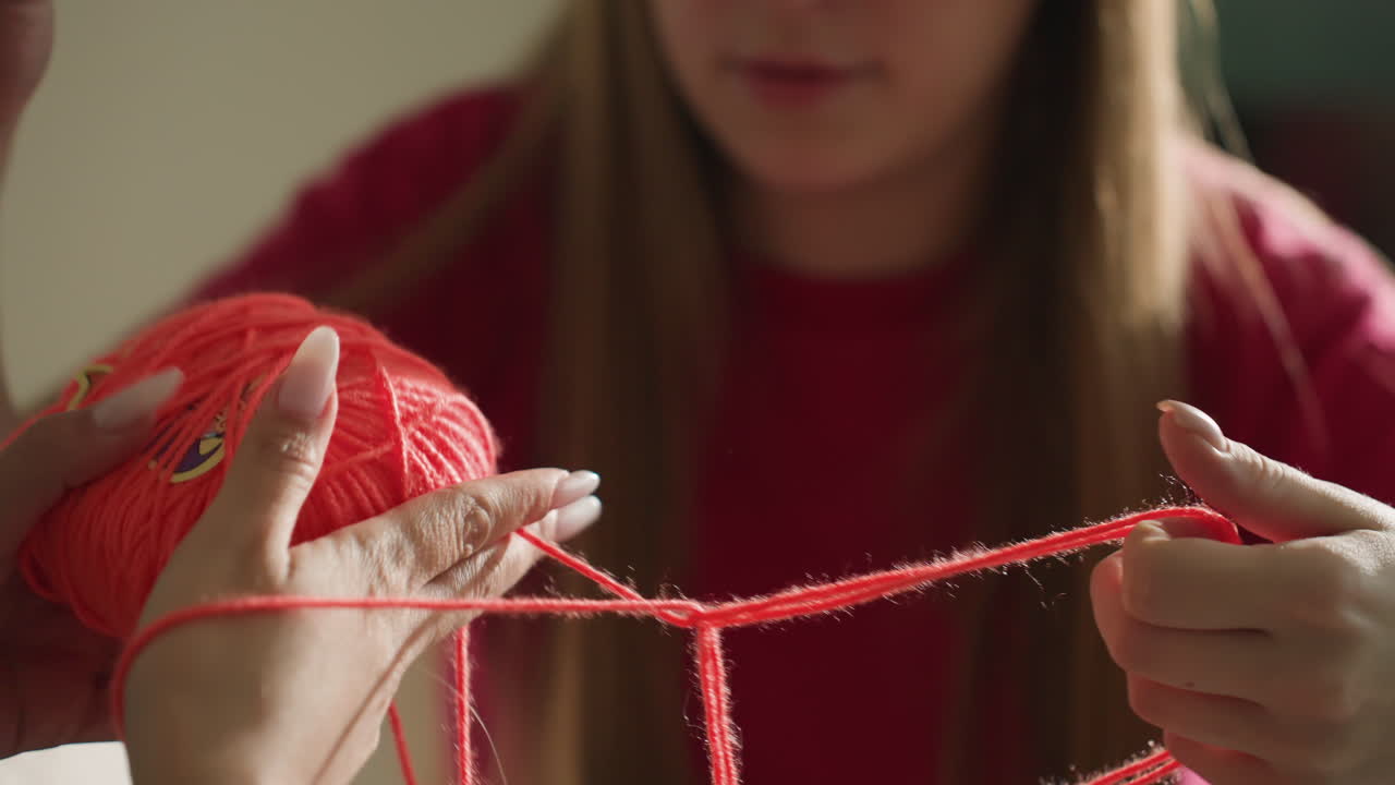 Amigas haciendo manualidades juntas, una joven aprende técnicas de artesanía, una amiga asiática enseña a una chica caucásica una técnica de nudos, un hilo rojo brillante está siendo cuidadosamente enlazado por las amigas en un acogedor ambiente interior.