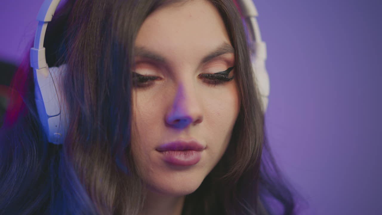 Close up young woman with long dark hair carefully putting on white headphones while surrounded by soft colorful lighting in cozy room, relaxed mood, fluffy sweater visible on arms