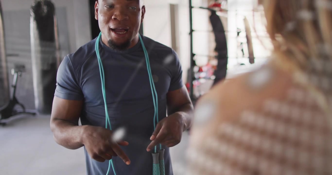 Trainer adjusting jump rope instructing client while gesturing down in gym as mirror overlay fading