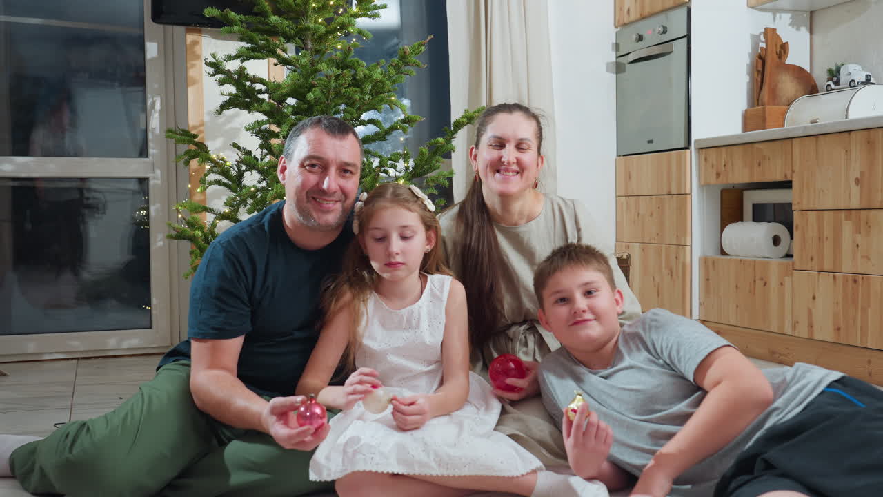Joyful family of four sitting closely together on soft white rug near decorated christmas tree in cozy modern kitchen holding ornaments during festive holiday season enjoying warm family moment
