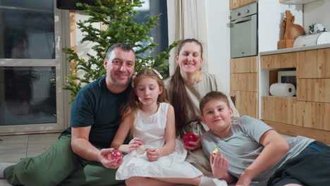 joyful family of four sitting closely together on soft white rug near decorated christmas tree in cozy modern kitchen holding ornaments during festive holiday season enjoying warm family moment