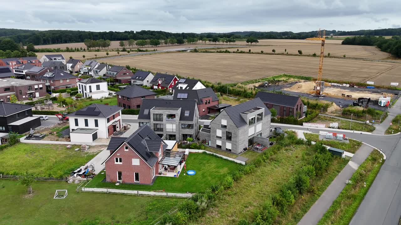 Aerial view of suburban housing development in Germany. Modern detached homes with gardens and gable roofs. Construction crane in background signals ongoing building activity. Farm fields in distance