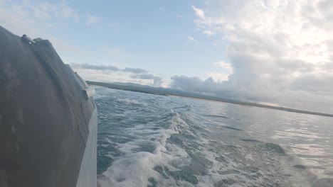 moving further away from an island of the galapagos islands. the boat is creating waves with its motion. white cloud and a blue sky are seen on the back