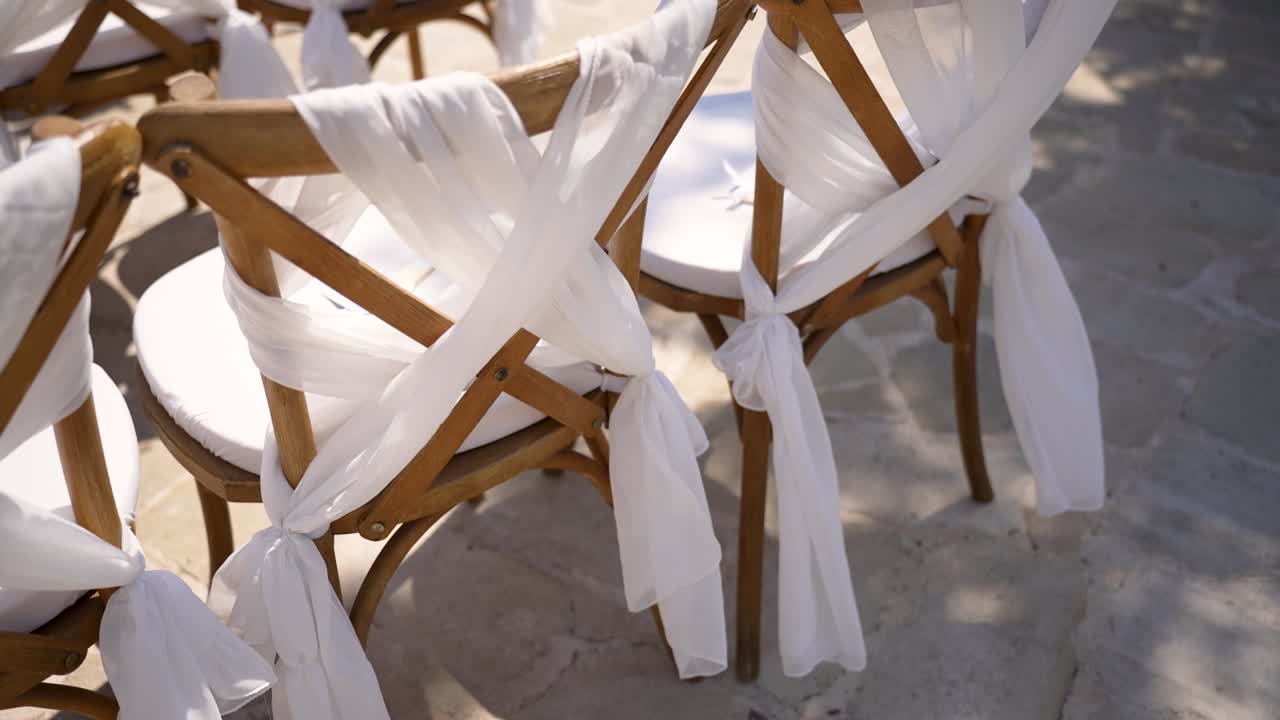 Beautifully arranged wooden chairs with soft white fabric ties create a welcoming atmosphere for an outdoor wedding. The sunny garden setting enhances the joyful occasion filled with love