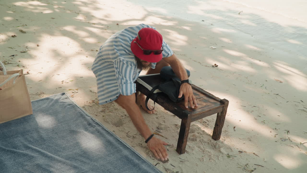 Uomo che prepara il tè in spiaggia con un piccolo sgabello di legno e una teiera, camicia a righe e berretto rosso, stuoia intrecciata sulla sabbia, luce solare a chiazze e ombra dagli alberi, mani che sistemano bollitore e vassoio, rituale intimo e calmo.