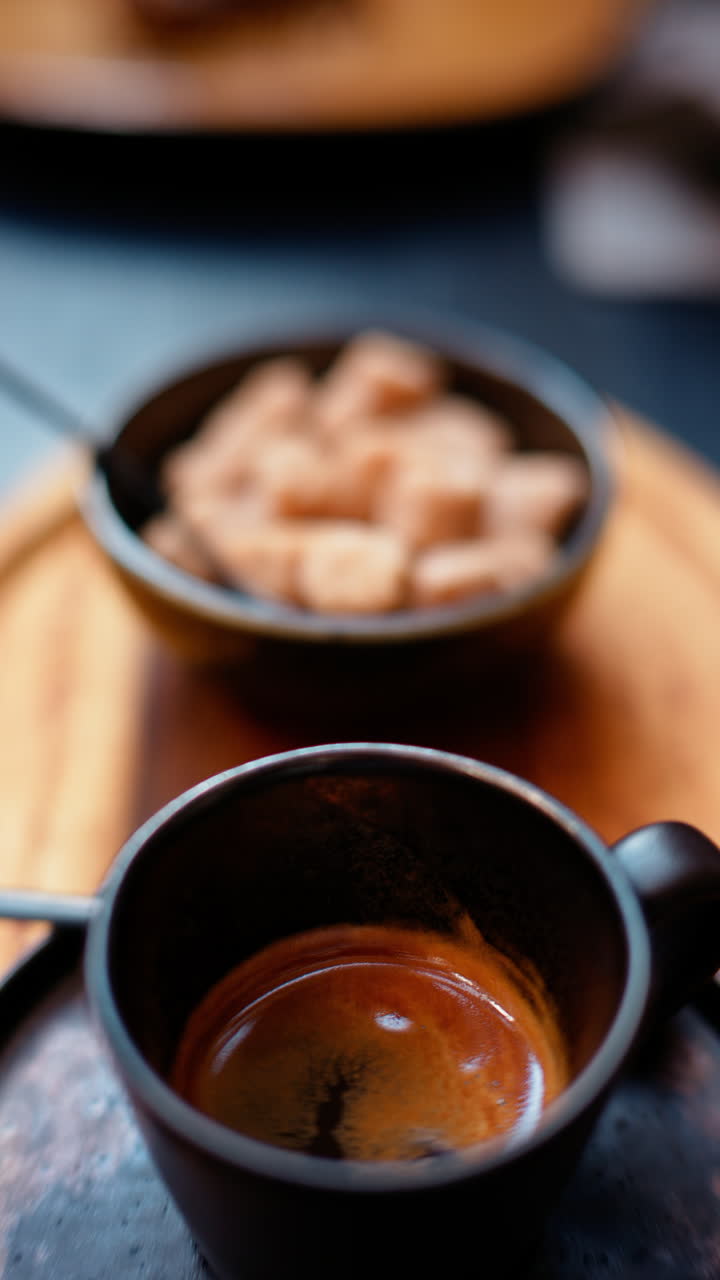 A black cup of espresso with a bowl of brown sugar cubes on a wooden tray. Vertical