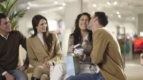 couples and friends sharing a joyful moment at a shopping mall, exchanging a gift and a kiss