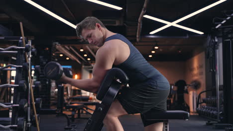 determined motivated sporty fit attractive caucasian male practicing weight training, performing single arm dumbbell bicep curls on preacher bench, workout in dark gym