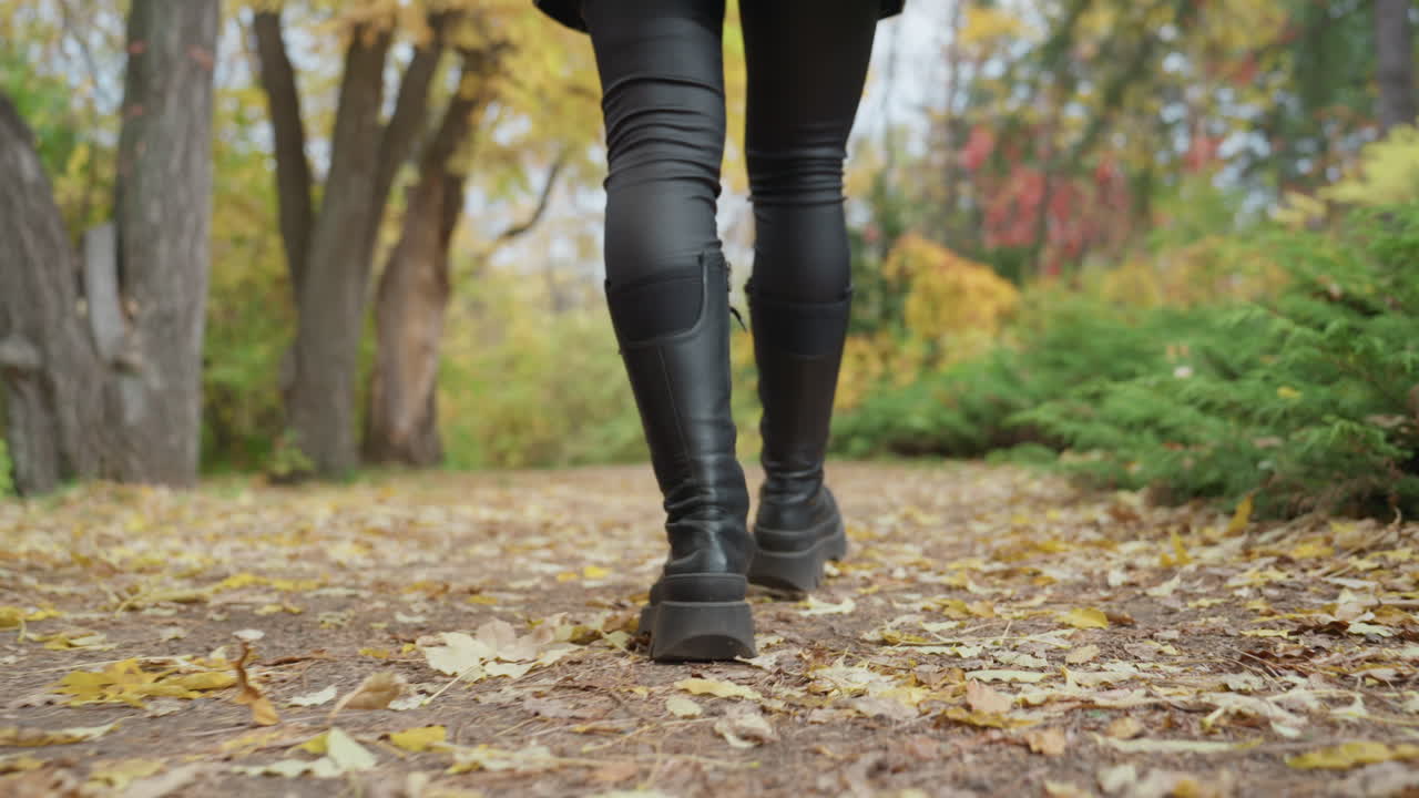 woman in leather boots walking leafstrewn park, closeup legs and boots, confident slow stride, leather pants with knee patches, crunchy fall leaves underfoot, golden foliage and soft bokeh, moody