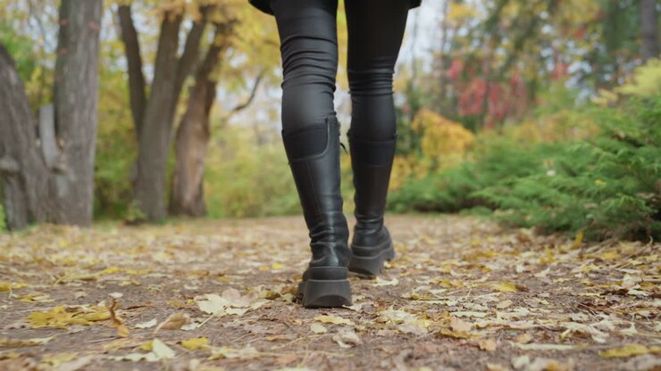 woman in leather boots walking leafstrewn park, closeup legs and boots, confident slow stride, leather pants with knee patches, crunchy fall leaves underfoot, golden foliage and soft bokeh, moody