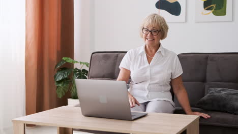 happy senior woman sitting on sofa in living room greeting and talking on video call on modern laptop 1