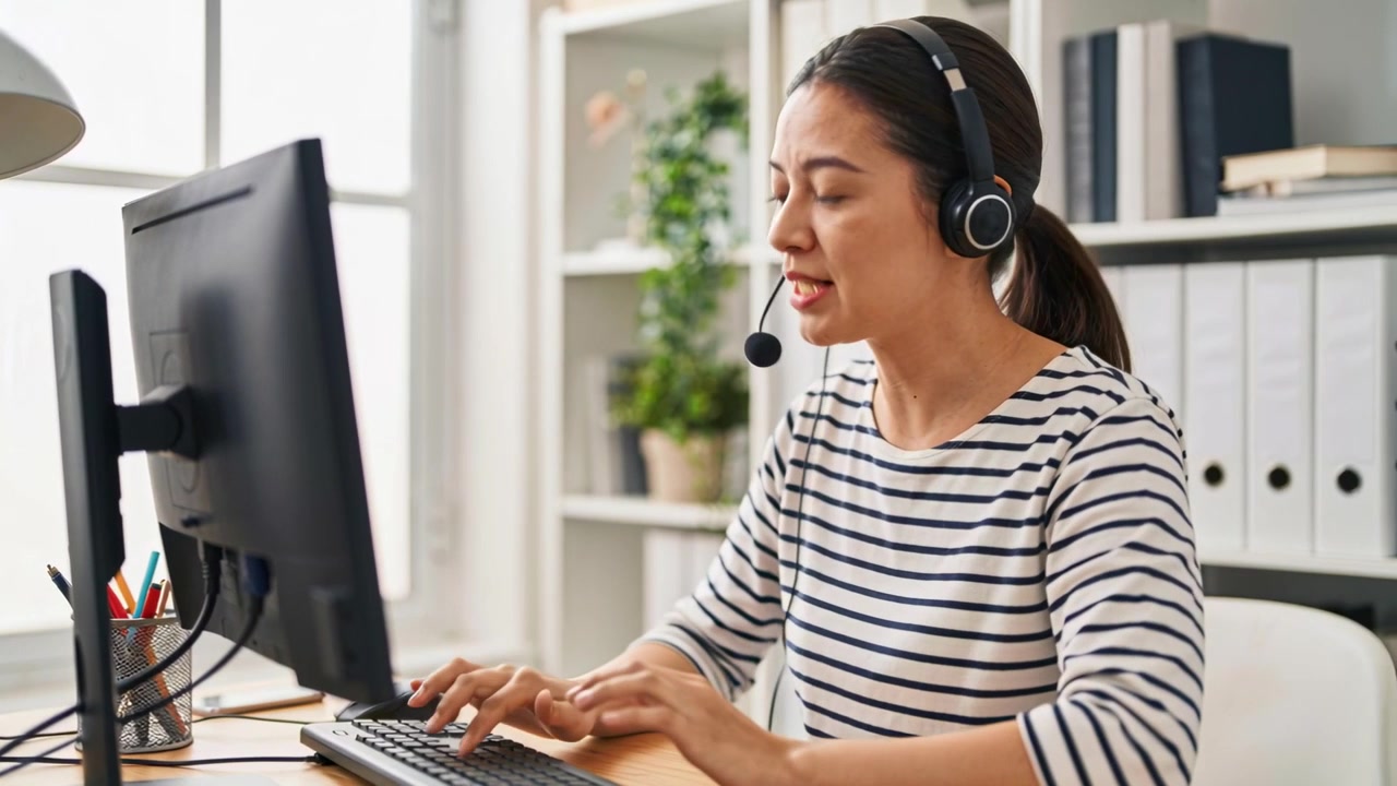 En asiatisk kvinne i stripete topp med hodesett starter en samtale ved pulten, gestikulerer og skriver notater.
