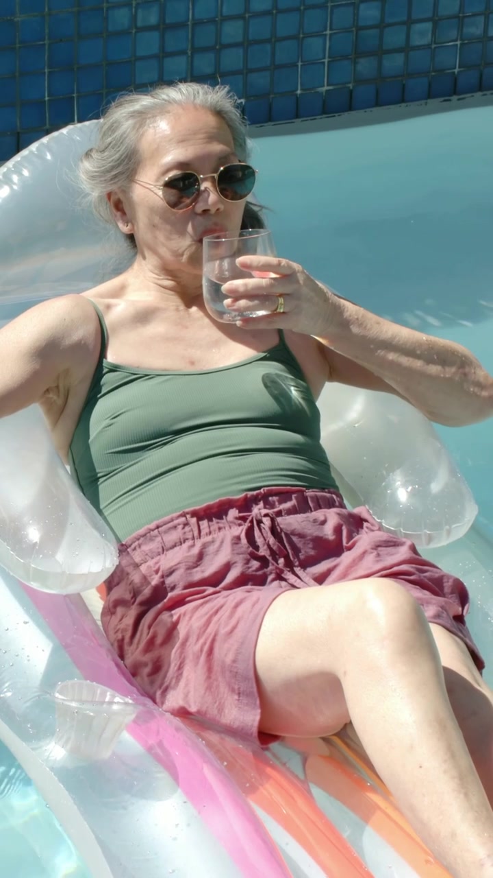Vertical video: Sun warming senior woman wearing green top sipping glass on float at pool cooling