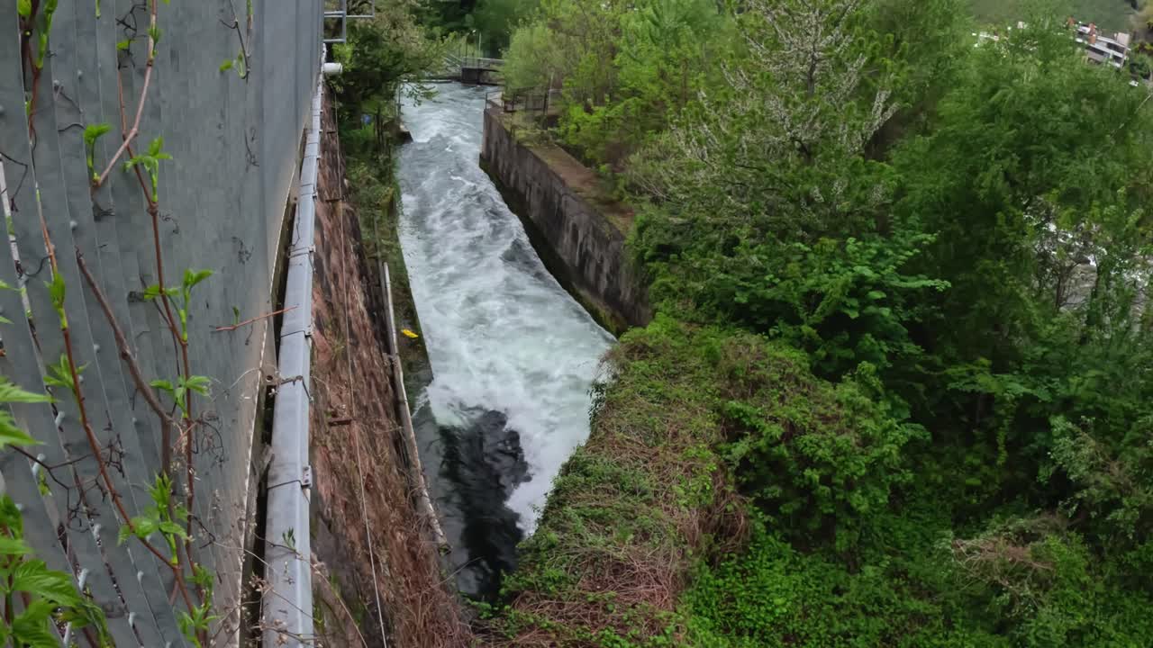 Above Lungotalvera in Bolzano, water flows through a dam toward Castel Roncolo.